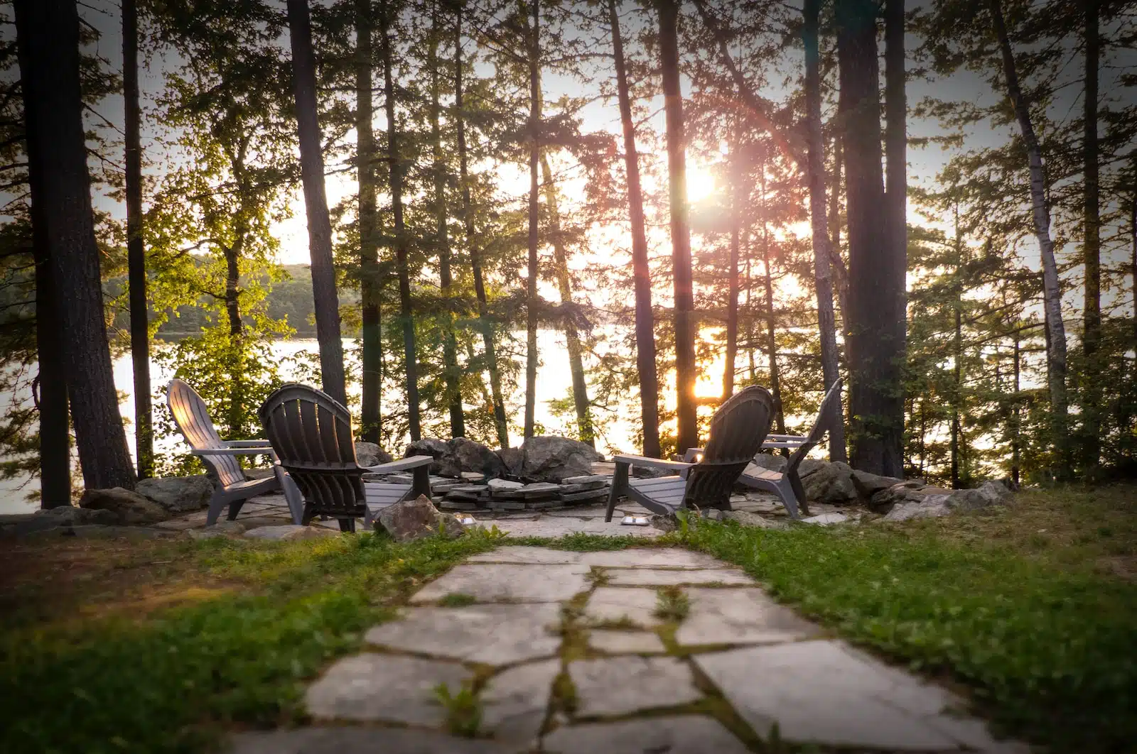 This image depicts a serene outdoor setting in a wooded area near a lake or body of water The scene features several Adirondack-style wooden chairs arranged on a stone patio or path surrounded by lush greenery and tall pine trees The warm golden light of the setting sun filters through the trees creating a peaceful and tranquil atmosphere The overall image conveys a sense of relaxation and connection with nature