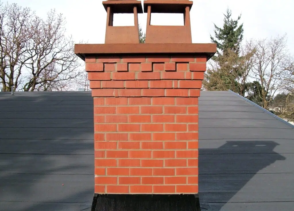 The image shows a brick chimney or fireplace structure with two flue openings at the top The chimney is constructed with red-orange bricks arranged in a uniform pattern The bricks have a slightly textured surface and are laid in a staggered pattern creating a visually interesting design The chimney is situated on top of a black or dark-colored roof which contrasts with the warm tones of the brickwork In the background there are bare trees suggesting a winter or early spring setting