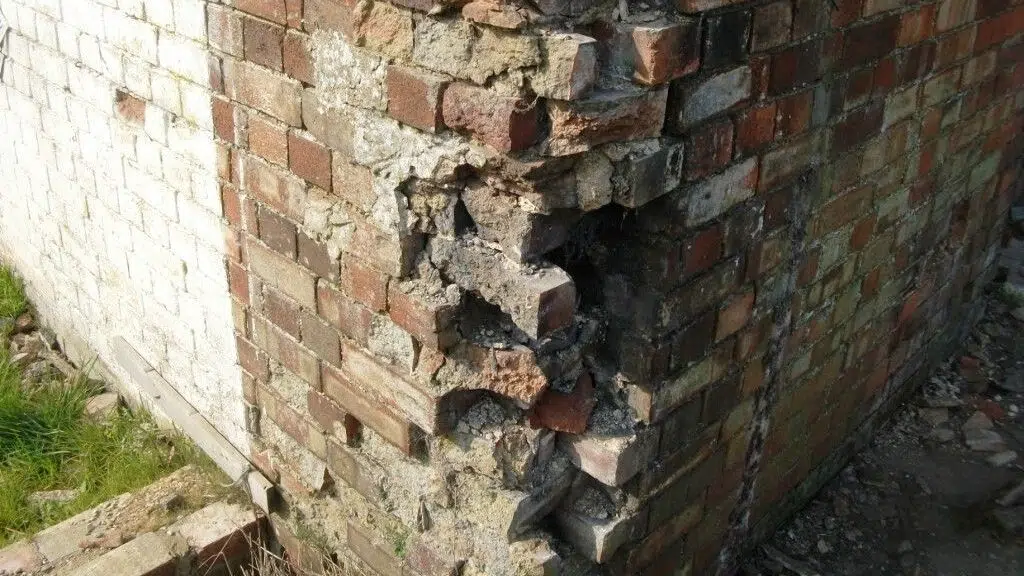 This image shows a section of a brick wall that has sustained some damage and deterioration The wall is composed of various sizes and shapes of bricks some of which appear to be crumbling or missing There are visible cracks and gaps in the mortar indicating the wall has experienced some structural issues over time The overall appearance suggests the wall is in need of repair or restoration to address the damaged areas