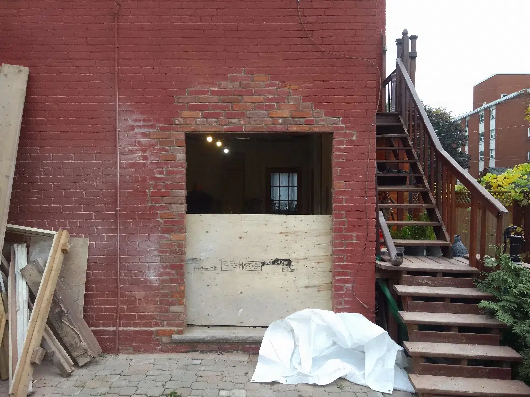 This image shows the exterior of a brick building with a recessed entryway The main wall is made of red bricks and there is a concrete or stone element in the entryway There is a wooden staircase leading up to a second-floor entrance and various construction materials and debris are visible in the foreground suggesting ongoing renovation or repair work The overall scene depicts a typical urban setting with a mix of architectural elements and signs of construction activity