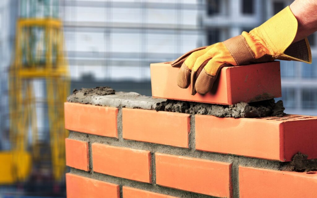 This image shows a close-up view of a construction workers hands wearing yellow protective gloves while laying bricks The bricks are a reddish-orange color and the worker is using a trowel to apply mortar to the bricks as they are being stacked to build a wall In the background there is a yellow construction scaffold visible indicating this is a construction site The overall image focuses on the hands and the process of brick laying highlighting the skilled work involved in masonry construction