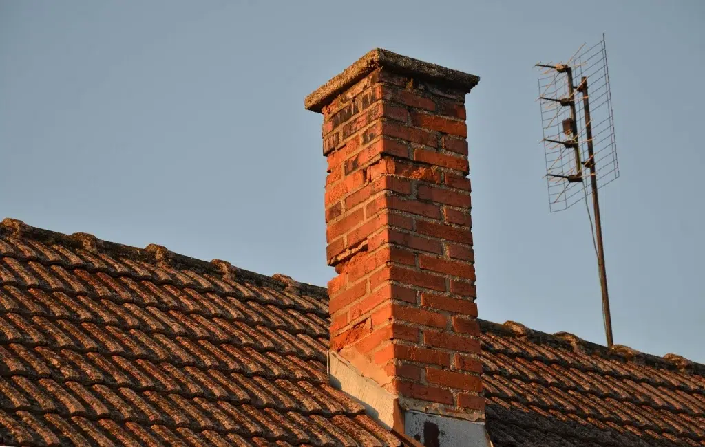 This image shows a brick chimney stack rising above a tiled roof The chimney is constructed of red bricks and has a distinctive square shape with a slightly tapered top The roof is made of dark weathered tiles that create a textured pattern In the background there is a metal structure possibly an antenna or satellite dish visible on the roof The overall scene depicts a traditional masonry structure and roofing elements commonly found in residential or commercial buildings