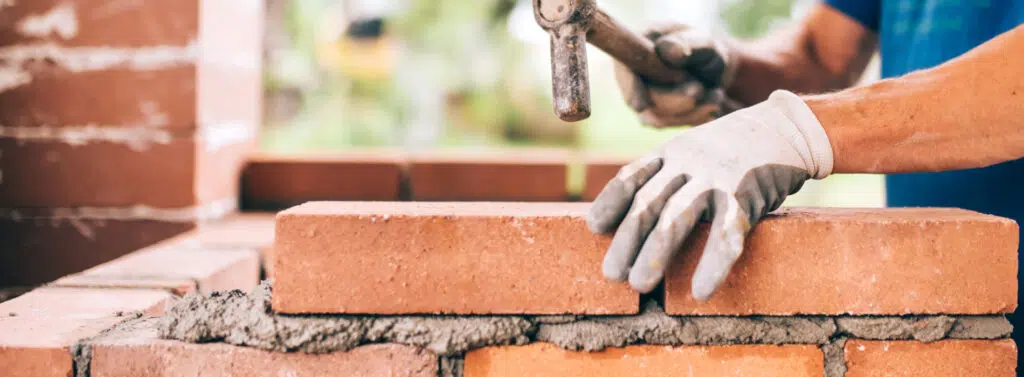 This image shows the hands of a construction worker wearing protective gloves laying bricks to build a wall The worker is using a hammer to carefully place a red brick onto the wall The background includes a partially constructed brick wall and a blurred natural setting suggesting an outdoor construction site The image highlights the skilled craftsmanship and manual labor involved in masonry work