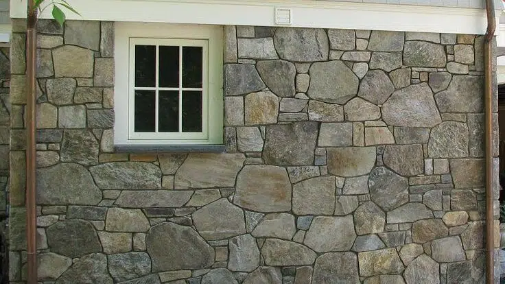 This image shows the exterior of a building with a stone wall facade The wall is constructed using various irregularly shaped and sized natural stone blocks creating a rustic textured appearance The stones are in shades of gray with some variations in color and texture A white-framed window is prominently featured in the center of the image providing a contrast to the rough stone surface The overall impression is of a sturdy well-crafted masonry construction