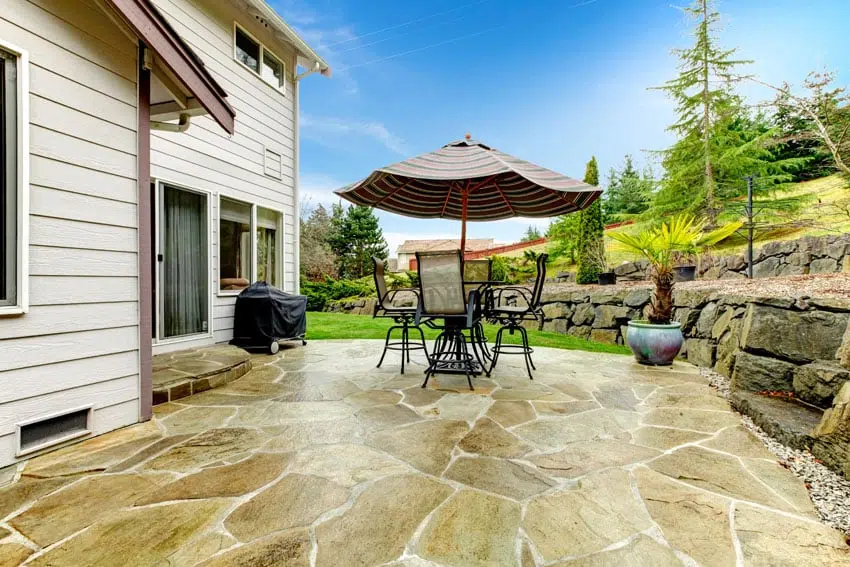 This image shows a cozy outdoor patio area behind a residential home The patio is made of natural stone pavers in shades of tan and brown creating an attractive and durable surface A large patio umbrella in a striped pattern provides shade over a small table and chairs creating a comfortable seating area The homes exterior is a light neutral color with wood accents and the surrounding landscape includes lush greenery such as trees and shrubs The overall scene conveys a peaceful and inviting backyard retreat