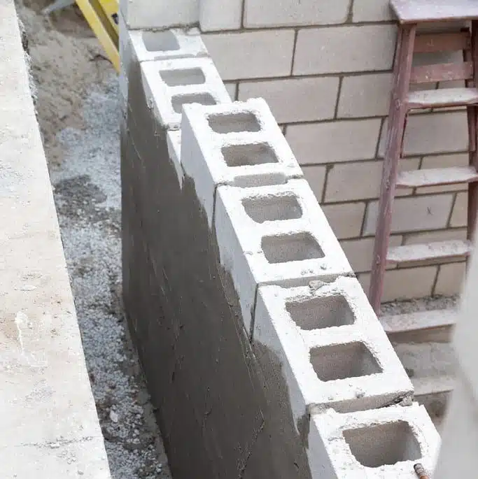 This image shows a stack of concrete blocks or cinder blocks The blocks are stacked against a brick wall with some debris or gravel visible at the base The blocks are gray in color and have a textured porous surface typical of concrete masonry units The image provides a close-up view of the stacked blocks highlighting their construction and placement in a masonry or construction setting
