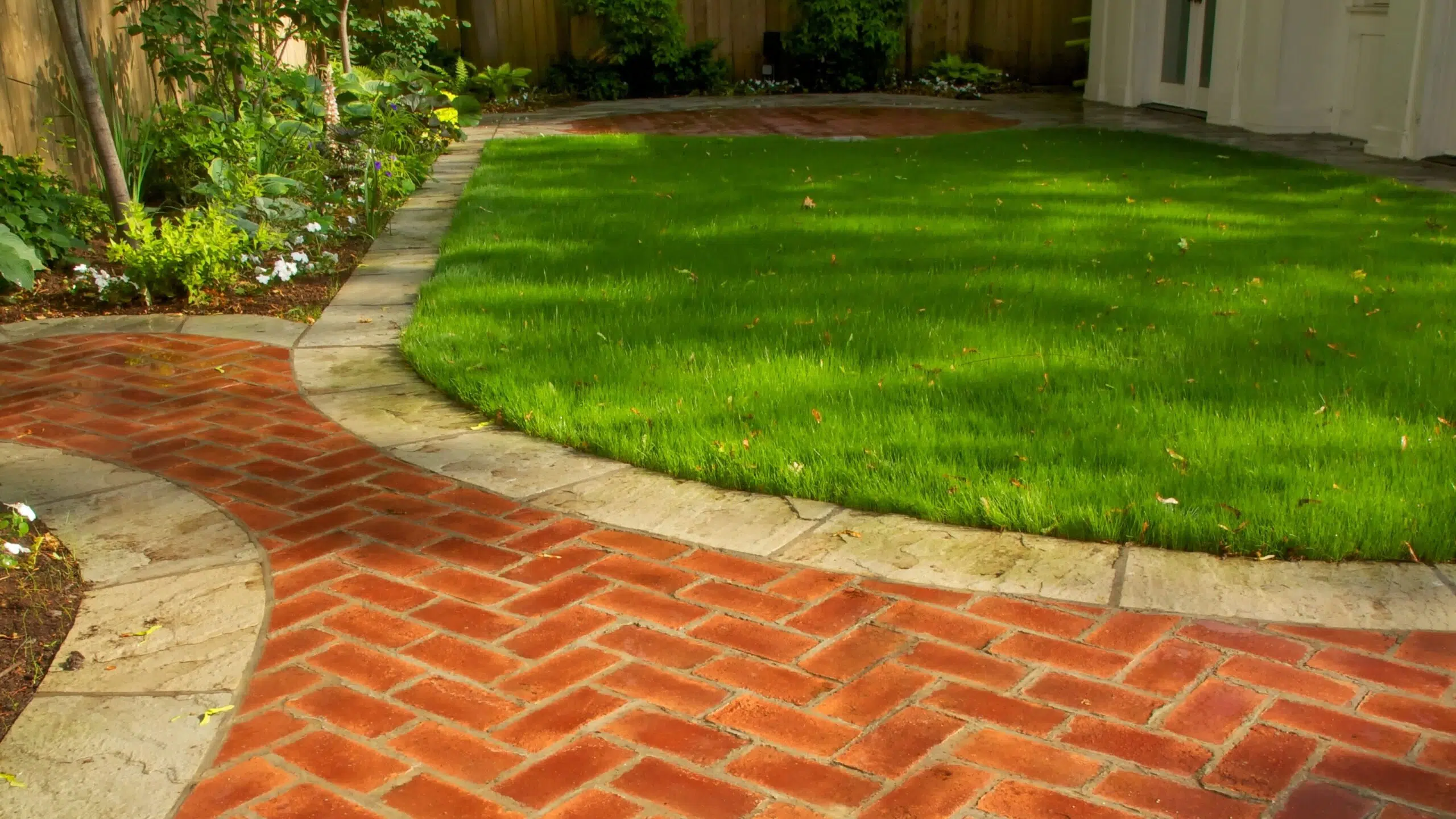 This image depicts a well-maintained backyard garden The focal point is a lush green lawn that covers the majority of the space Surrounding the lawn is a brick pathway that winds through the garden leading to a white building in the background The brick pathway has a herringbone pattern adding visual interest to the design Alongside the path there are various plants and shrubs creating a naturalistic and inviting atmosphere The overall scene conveys a sense of tranquility and attention to detail in the landscaping