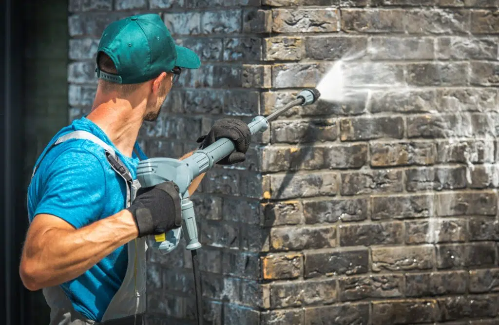 
An image shows a person using a pressure washer to clean a brick wall. The individual is wearing a blue shirt and a cap, focusing the high-pressure water stream on the dark brick surface to remove dirt and grime. The spray creates a visible area of lighter brick, indicating the cleaning process's effectiveness.