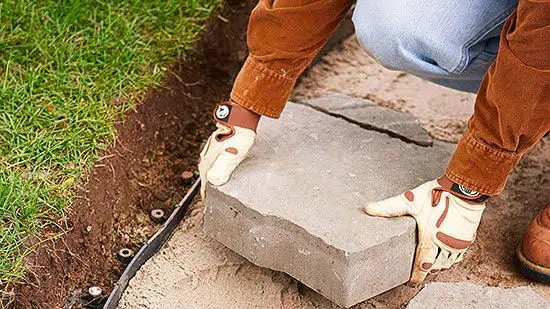 
A person wearing gloves is installing a large square paving stone onto a prepared sand base, with the edge of the lawn and garden edging visible nearby. The individual is in a crouched position, focused on carefully placing the stone in the correct position for a pathway or patio. Attention to detail is evident in the careful alignment of the paving stone with the existing edge.
