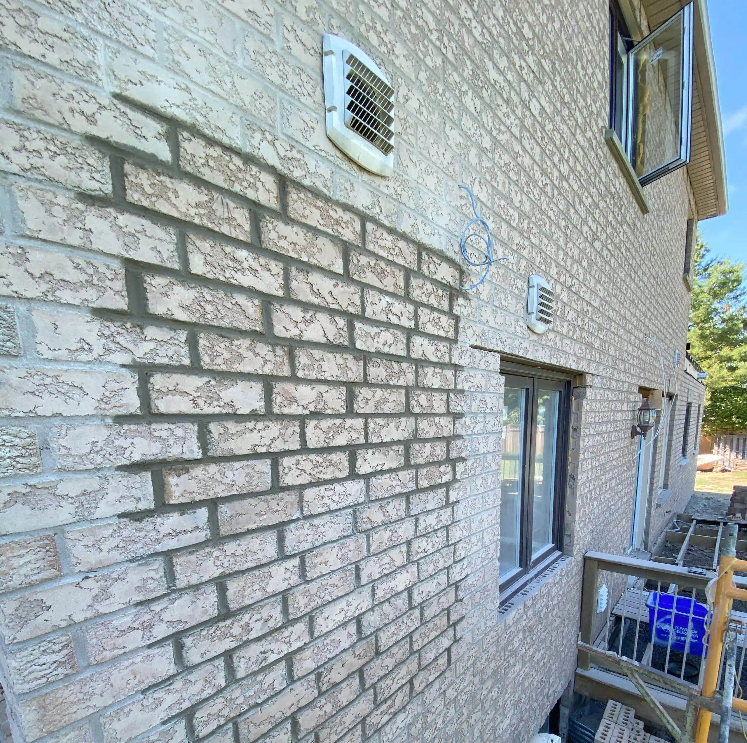 This image shows the exterior wall of a building constructed with light-colored brick The bricks have a textured weathered appearance creating an interesting visual pattern on the wall There is a small ventilation grill or vent installed in the wall and a window is visible on the right side of the image The overall scene suggests a residential or commercial building exterior
