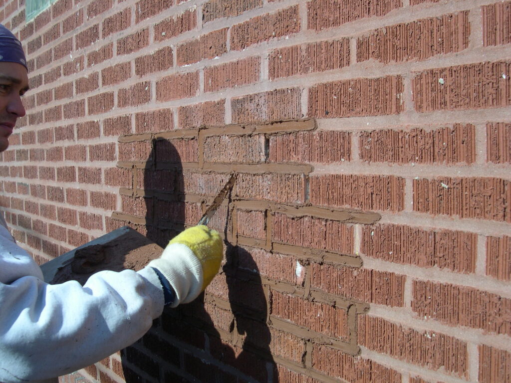 The image shows a close-up view of a brick wall A persons hand wearing a yellow glove is visible holding a tool and working on repairing or fixing a damaged section of the brick wall The bricks appear to be a reddish-brown color and have a textured irregular pattern The overall scene depicts a masonry repair or restoration work being carried out on the brick wall