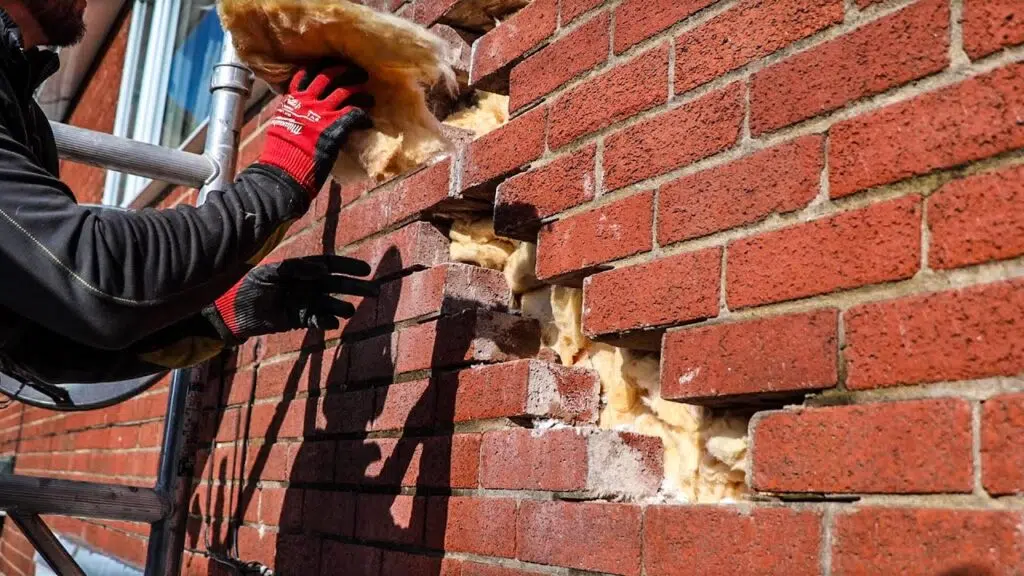 This image shows a close-up view of a persons hands wearing red protective gloves while repairing a brick wall The person is using a tool likely a chisel or hammer to remove damaged or crumbling bricks from the wall The wall is made of reddish-brown bricks and the image captures the textures and patterns of the brickwork The persons hands and the tool they are using are the main focus of the image highlighting the manual labor and skill involved in masonry repair work