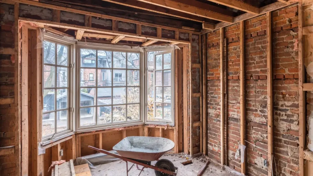 The image shows the interior of a partially renovated room The walls are made of exposed brick and wooden beams are visible in the ceiling There is a large window with multiple panes that provides natural light into the space In the foreground there is a wheelbarrow and some construction materials indicating that this room is currently undergoing renovation or remodeling work