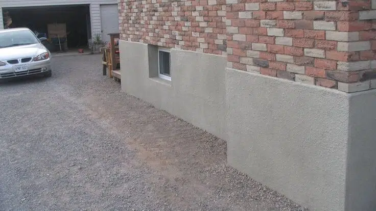 A residential driveway alongside a house showcasing a contrast between the upper brick wall and the lower section with a smooth parging finish including two small basement windows The parged area provides a neat protective coating against the foundation of the home