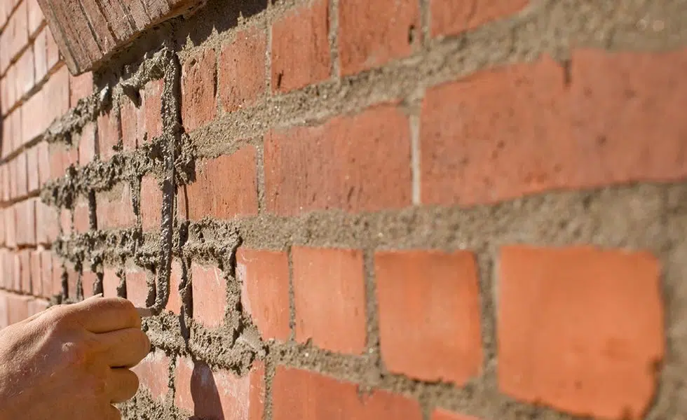 This image shows a close-up view of a brick wall The bricks are a reddish-orange color and the mortar between them is a light gray The bricks appear to be laid in a standard running bond pattern with each brick overlapping the joints between the bricks in the row below A hand is visible in the foreground pointing to or touching one of the bricks indicating that this may be an image related to construction or masonry work
