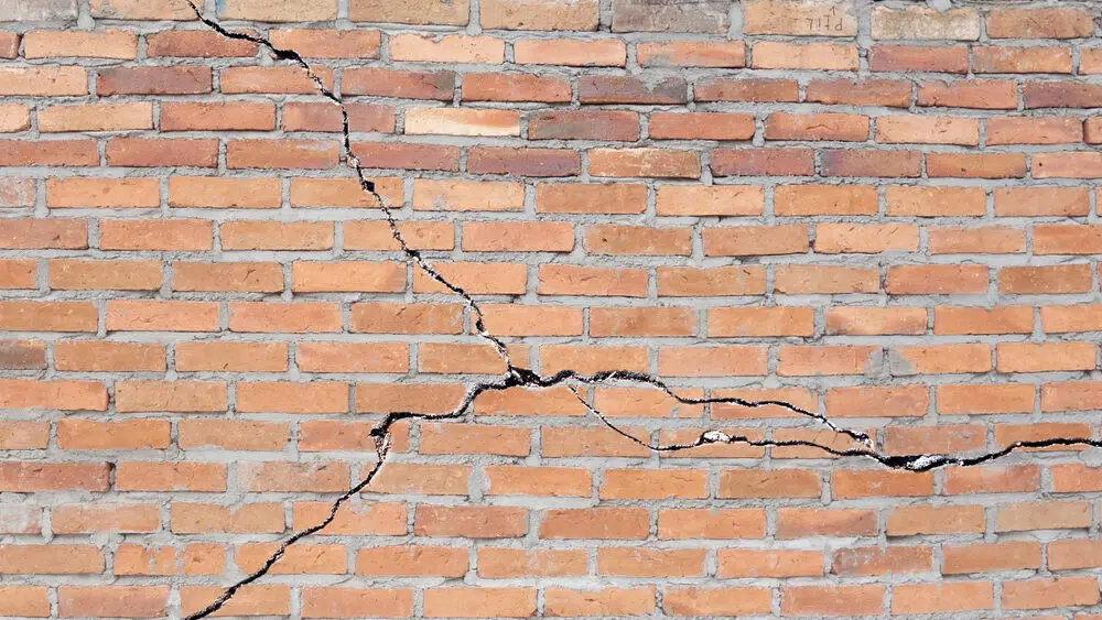 This image shows a cracked and damaged brick wall The wall is composed of rows of orange-colored bricks with several visible cracks running horizontally and vertically across the surface The cracks appear to be significant indicating potential structural issues or weathering damage to the wall over time The overall texture and pattern of the bricks are clearly visible providing a detailed view of the masonry construction