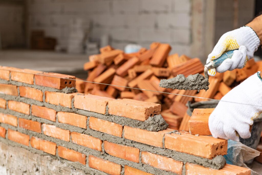 This image shows a close-up view of a bricklaying process It depicts a persons hands wearing white gloves laying bricks and applying mortar to construct a brick wall The bricks are a reddish-orange color and the mortar used to bind them together is a grayish-white cement-based material In the background there are additional bricks and construction materials indicating an ongoing masonry project