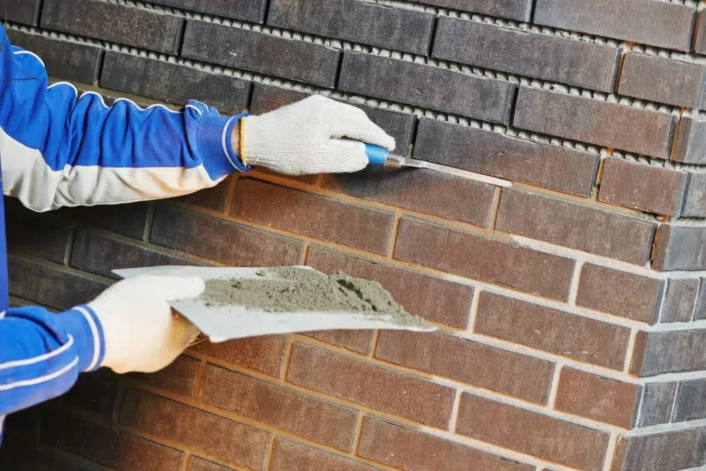 This image shows a persons hands wearing blue and white work gloves while applying mortar or grout to a brick wall The person is using a trowel to spread the mortar evenly between the bricks which have a dark textured appearance The background consists of additional rows of bricks indicating this is a construction or masonry work site