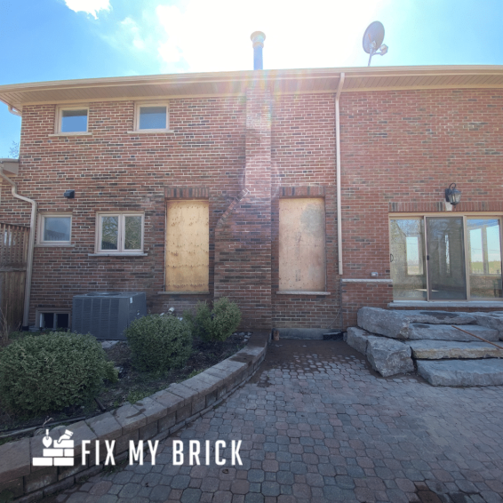 This image shows the exterior of a brick building belonging to the company "Fix My Brick". The building has a brick facade with several windows and two boarded-up doorways. There is a paved walkway leading to the entrance, with decorative stone accents and landscaping around the building. The image conveys a sense of a commercial or industrial property in need of repair or renovation services provided by Fix My Brick.